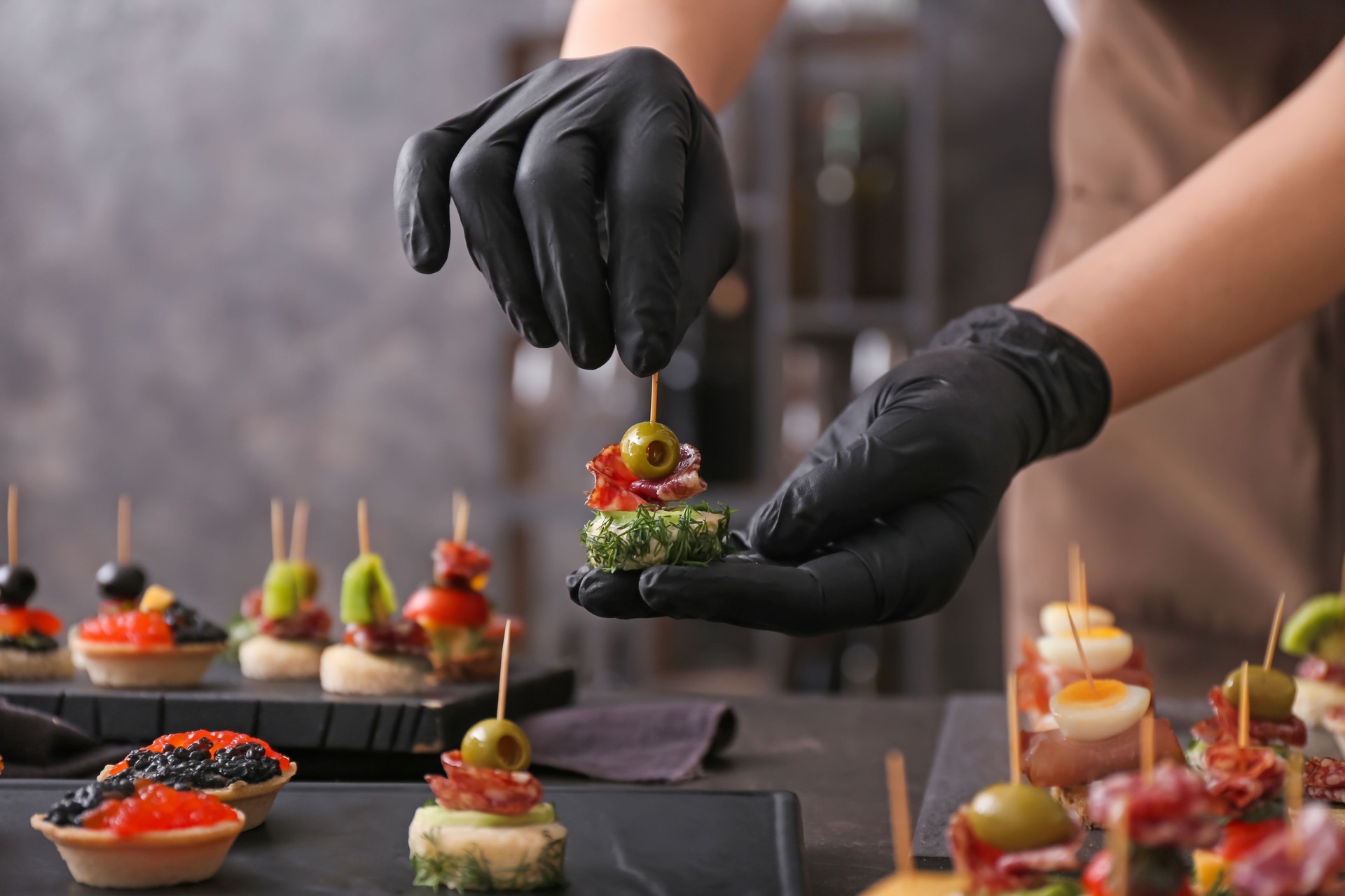 Professional caterer wearing black gloves carefully arranging and garnishing savory canap?s and appetizers on a dark platter during an upscale event.